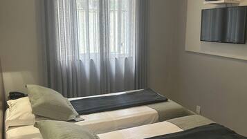 Una habitación de hotel con dos camas, una ventana con cortinas translúcidas, un televisor de pantalla plana en la pared y un termostato sobre el televisor.