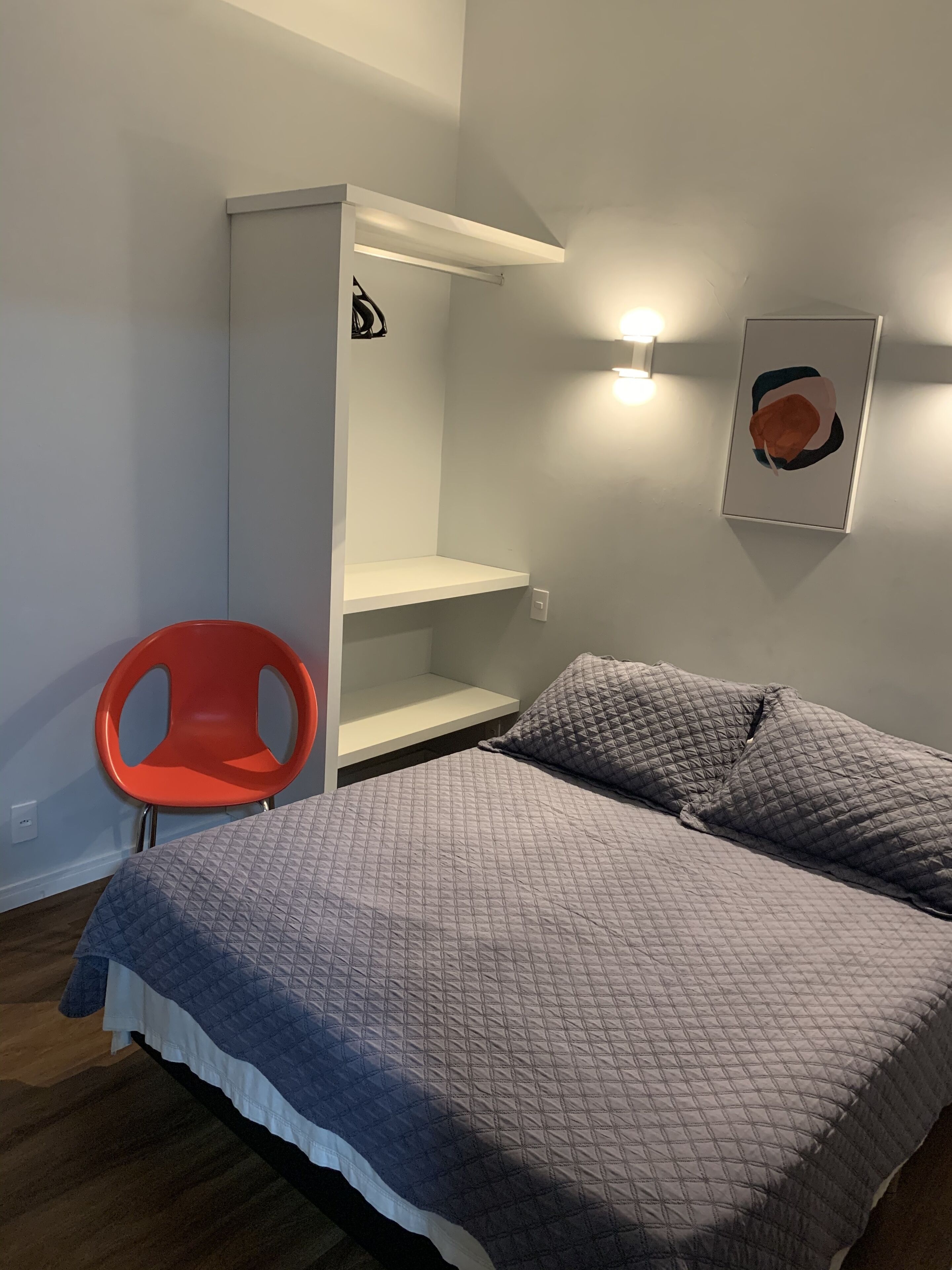 A bedroom with a bed, a chair, a shelf unit, and a wall-mounted light fixture.
