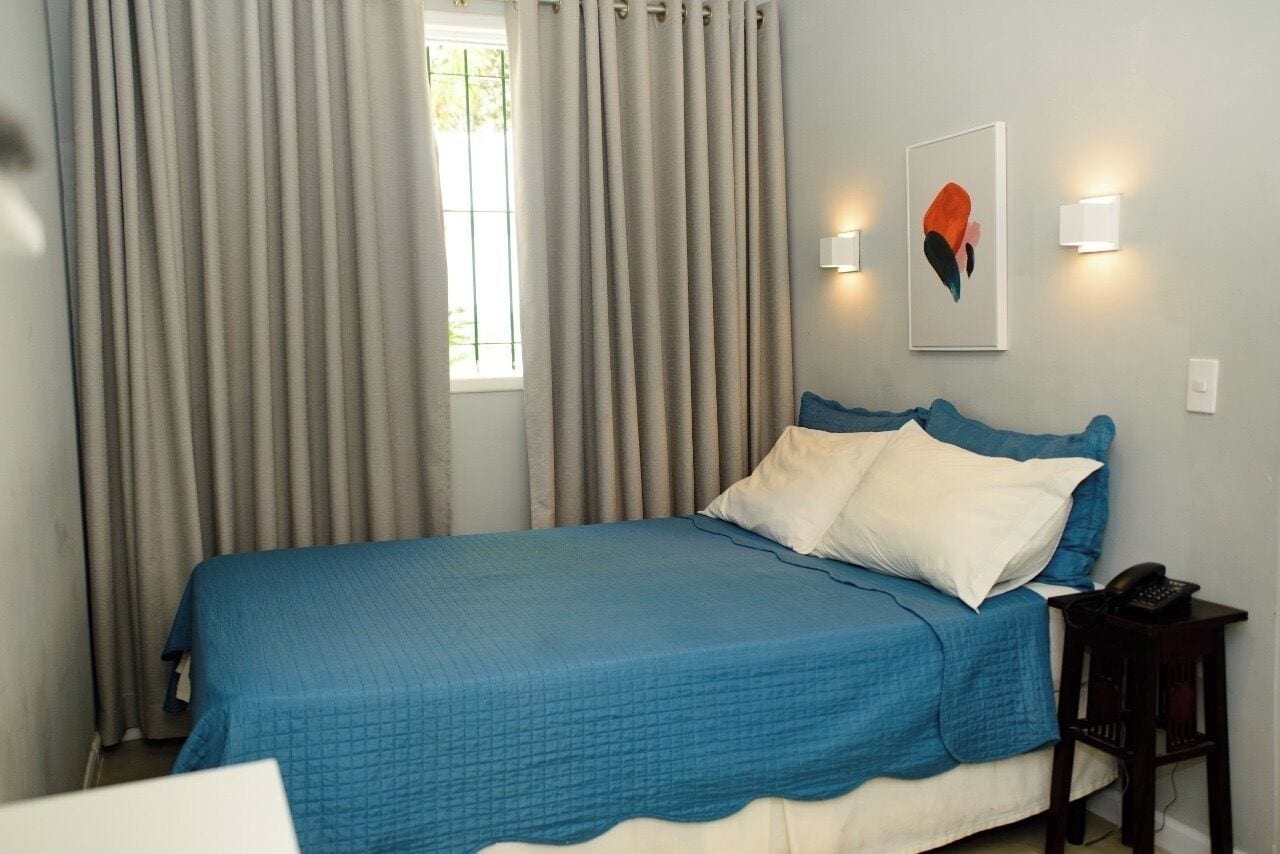 A neatly made bed with a blue bedspread and white pillows, a bedside table with a phone, and a window with curtains.