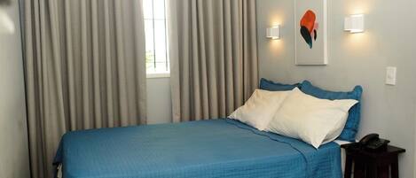 Un letto ben rifatto con copriletto blu e cuscini bianchi, un comodino con un telefono e una finestra con tende.