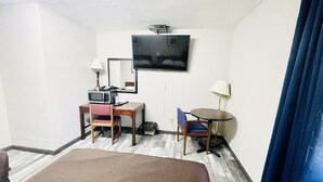 A hotel room with a bed, a desk with a computer, a chair, a small table, a TV, and a mirror.