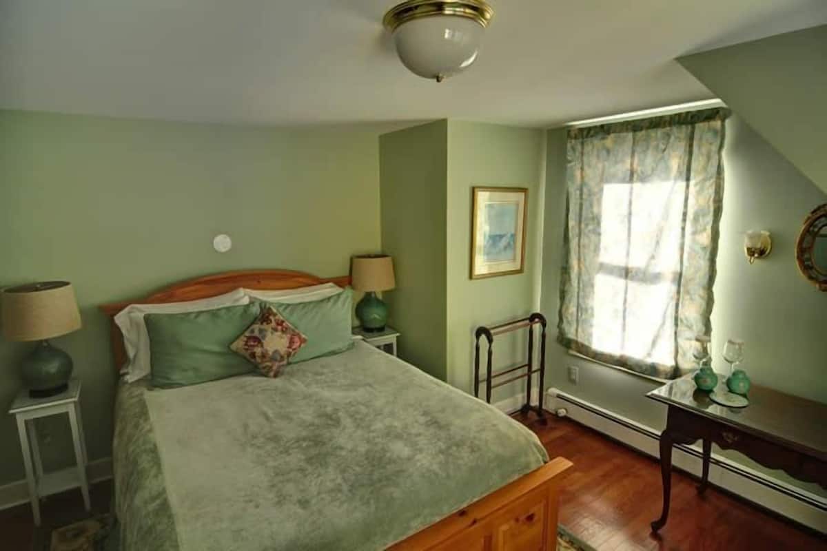 A bedroom with a bed, two bedside tables, a nightstand, a window with curtains, a picture on the wall, and a ceiling light fixture.