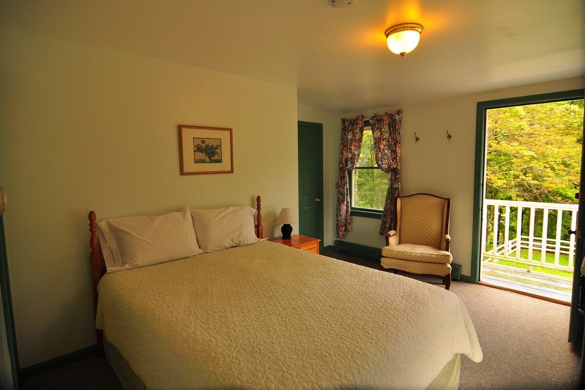 A bedroom with a large bed, a chair, a window with curtains, and a view of greenery outside.