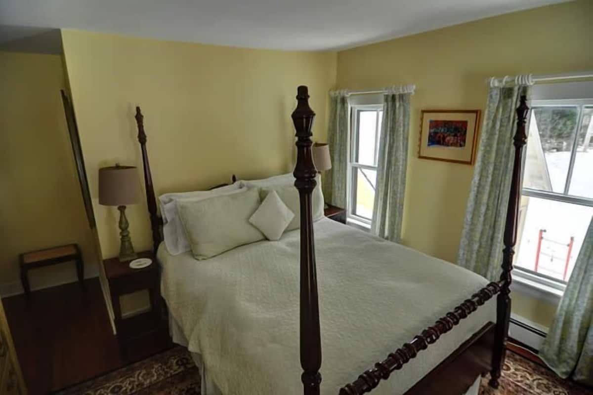 A bedroom with a four-poster bed, a bedside table with a lamp, a window with curtains, and a framed picture on the wall.