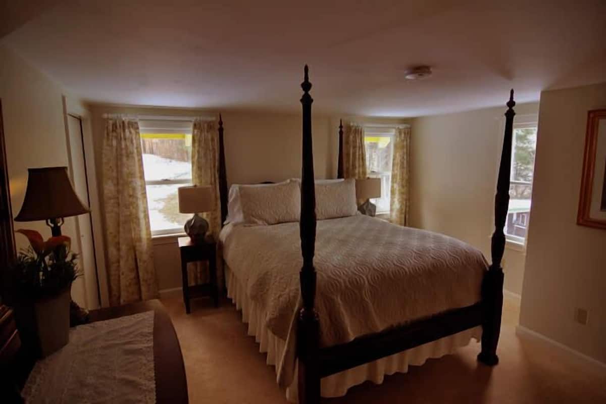 A four-poster bed with a white bedspread, a bedside table with a lamp, and a window with curtains.