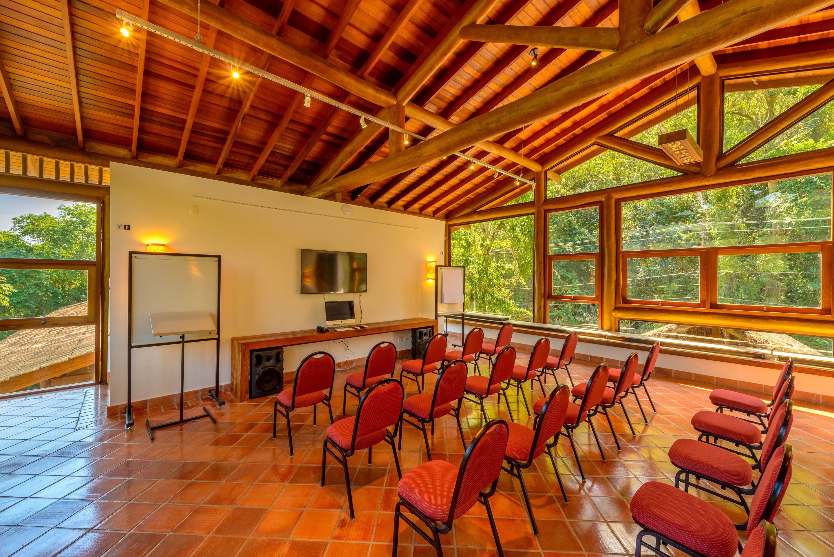 Un salón de conferencias con vigas de madera, sillas rojas, una pizarra y ventanas amplias.