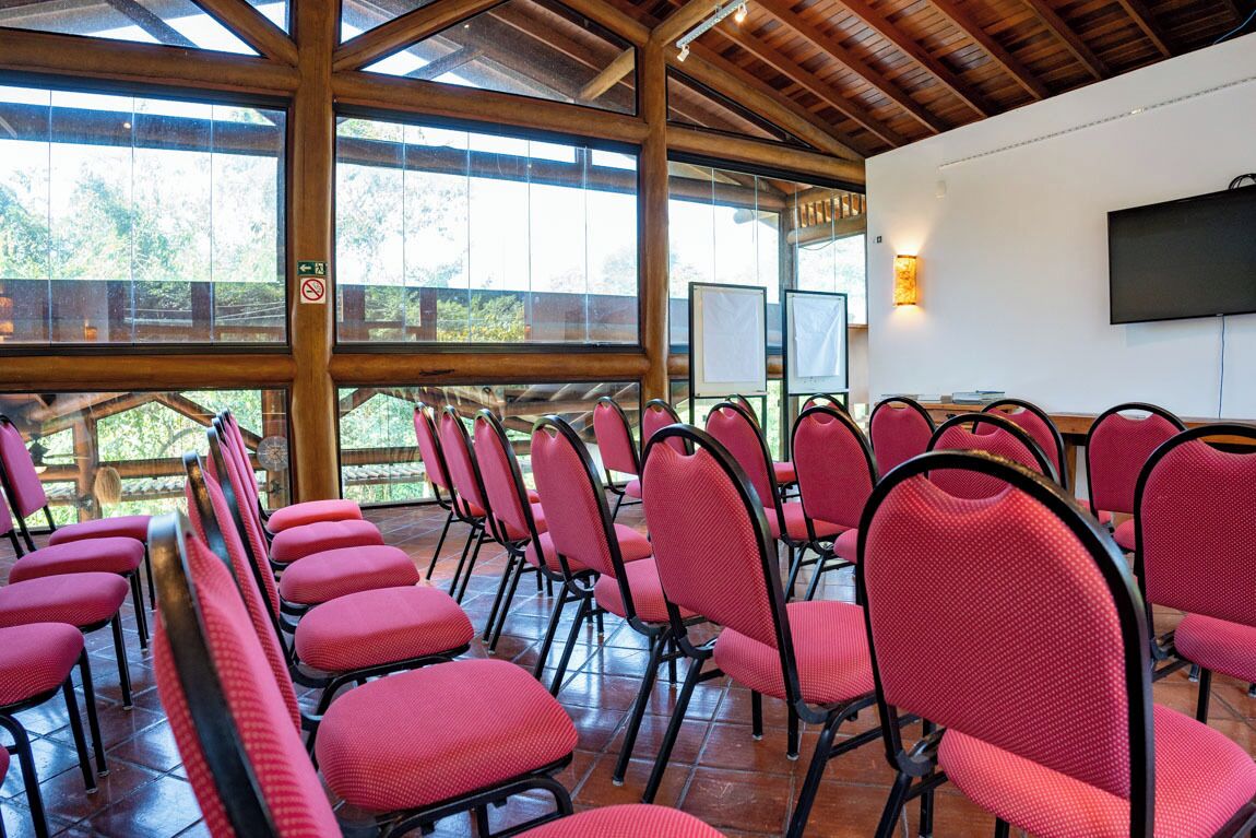 Un salón de conferencias con hileras de sillas rojas, vigas de madera, ventanas amplias y un televisor montado en la pared.