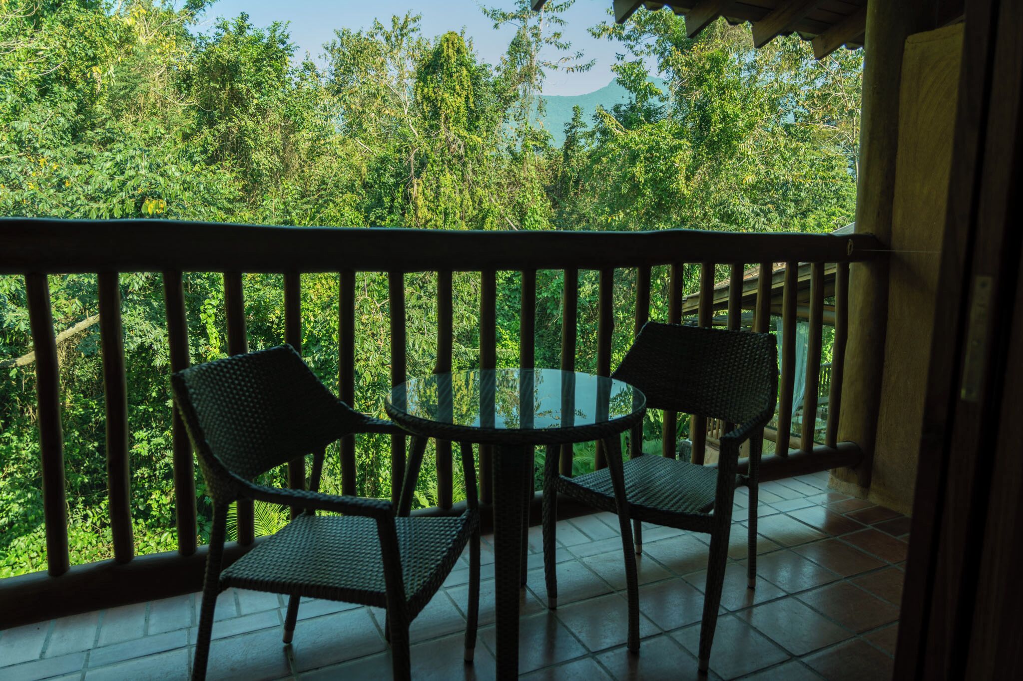 A balcony with two wicker chairs and a glass table, overlooking a lush green landscape.