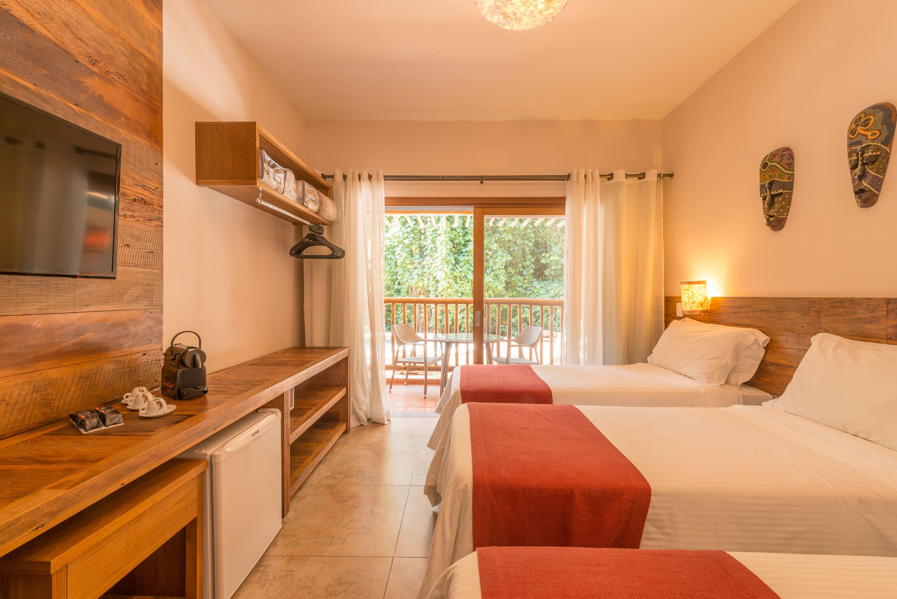 A hotel room with a wooden desk, a television, two beds with red bedspreads, a small refrigerator, a balcony with a table and chairs, and a large window with curtains.