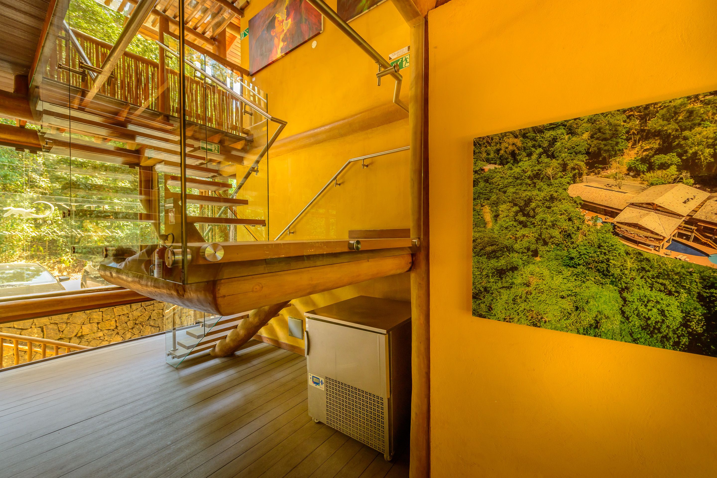 A wooden staircase with a glass balustrade leading to a balcony, a small air conditioning unit, and a framed picture of a vehicle on the wall.