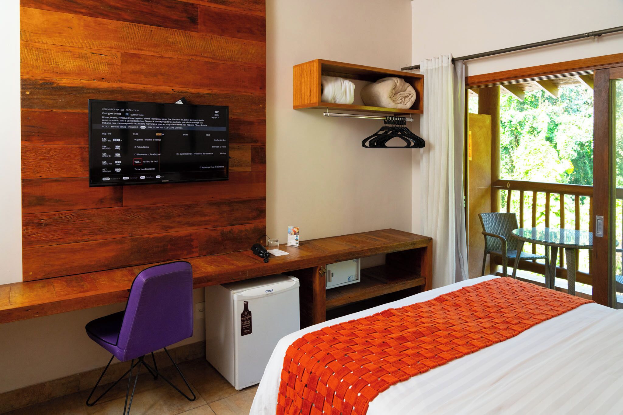 Een hotelkamer met een houten bureau, een kleine koelkast, een bed met een oranje deken, een aan de muur bevestigde televisie en een raam met uitzicht op groen.