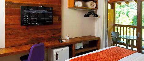 A hotel room with a wooden desk, a small refrigerator, a bed with an orange blanket, a TV mounted on the wall, and a window with a view of greenery.