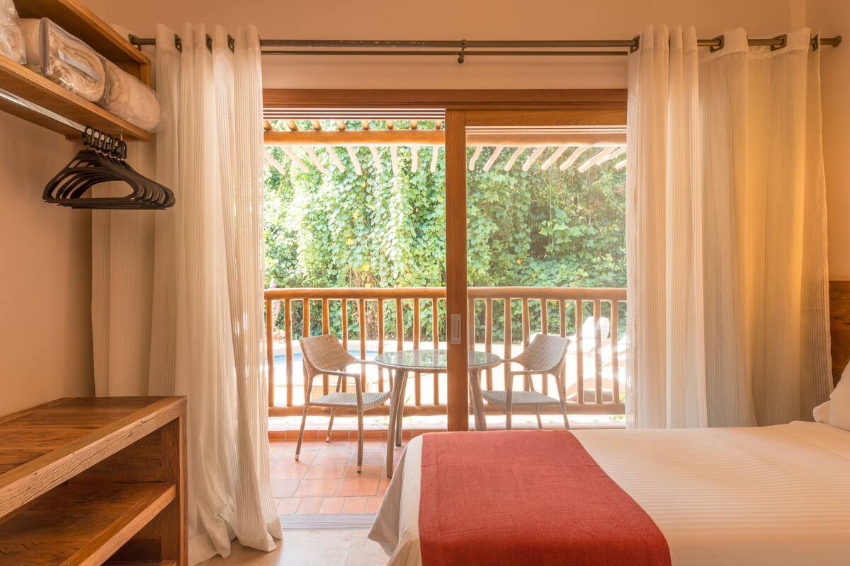 A bedroom with a bed, a wooden shelf, a hanging rack, and a balcony with chairs and a table.