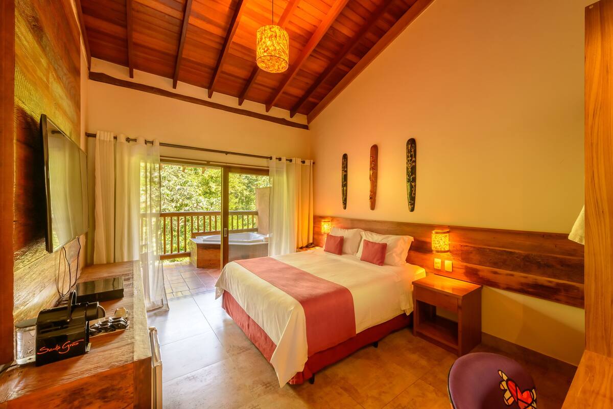 A spacious bedroom with a large bed, a wooden ceiling, a balcony with a view of greenery, and a TV on a wooden stand.