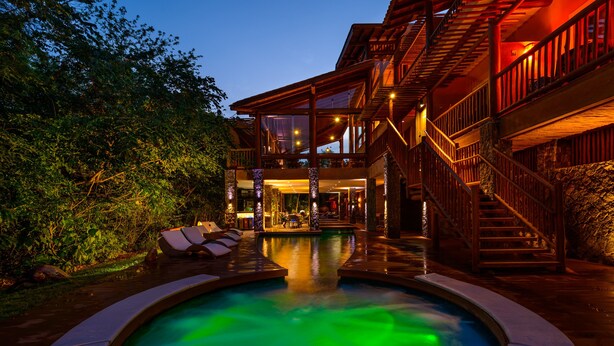 Une piscine dotée d’une terrasse en bois et de chaises longues, entourée d’un mur de pierre et d’une végétation luxuriante.