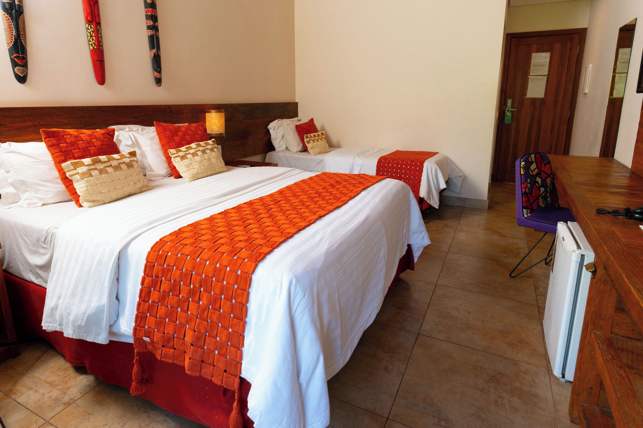 Habitación de hotel con dos camas, cabecero de madera, una mesita, una silla y un refrigerador.