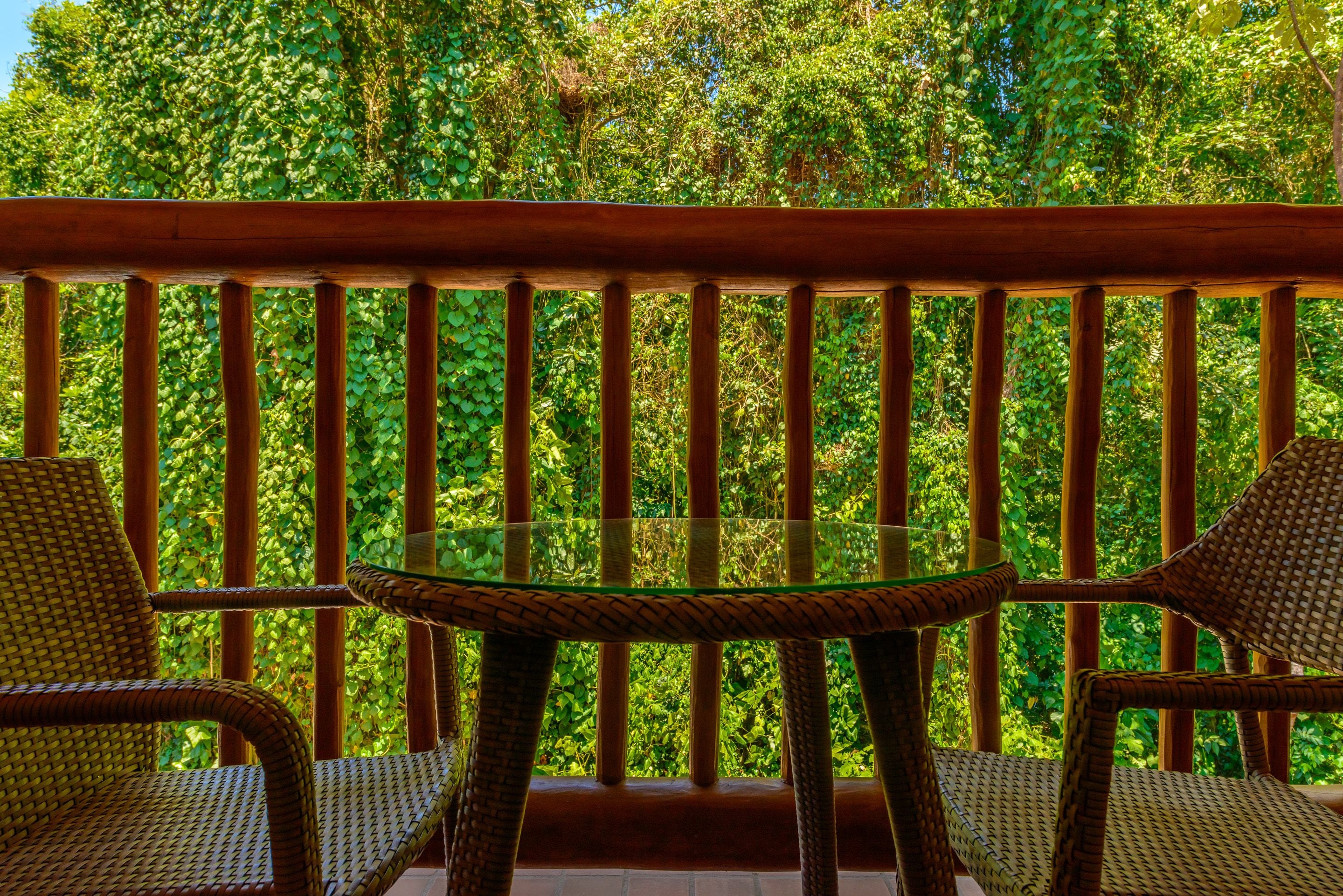 A wooden balcony with wicker chairs and a glass table, surrounded by lush green foliage.
