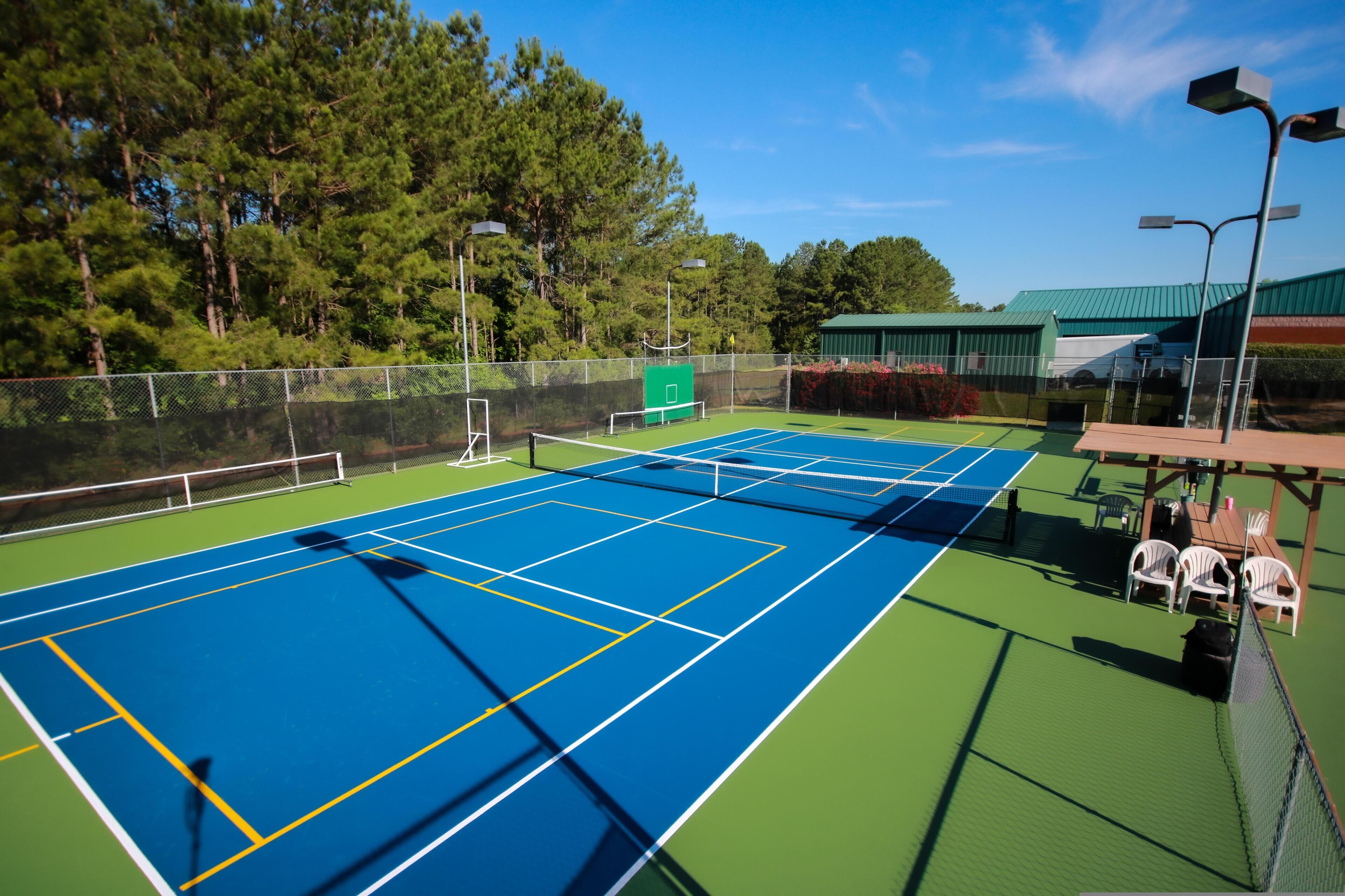 A tennis court with multiple courts, surrounded by a fence, and a seating area with tables and chairs.