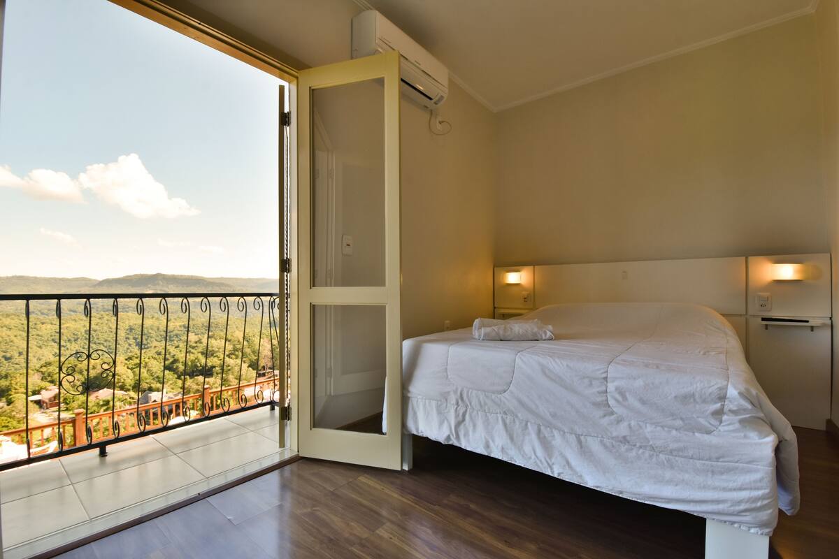 A bedroom with a large bed, an open sliding door to a balcony, and a view of a mountainous landscape.