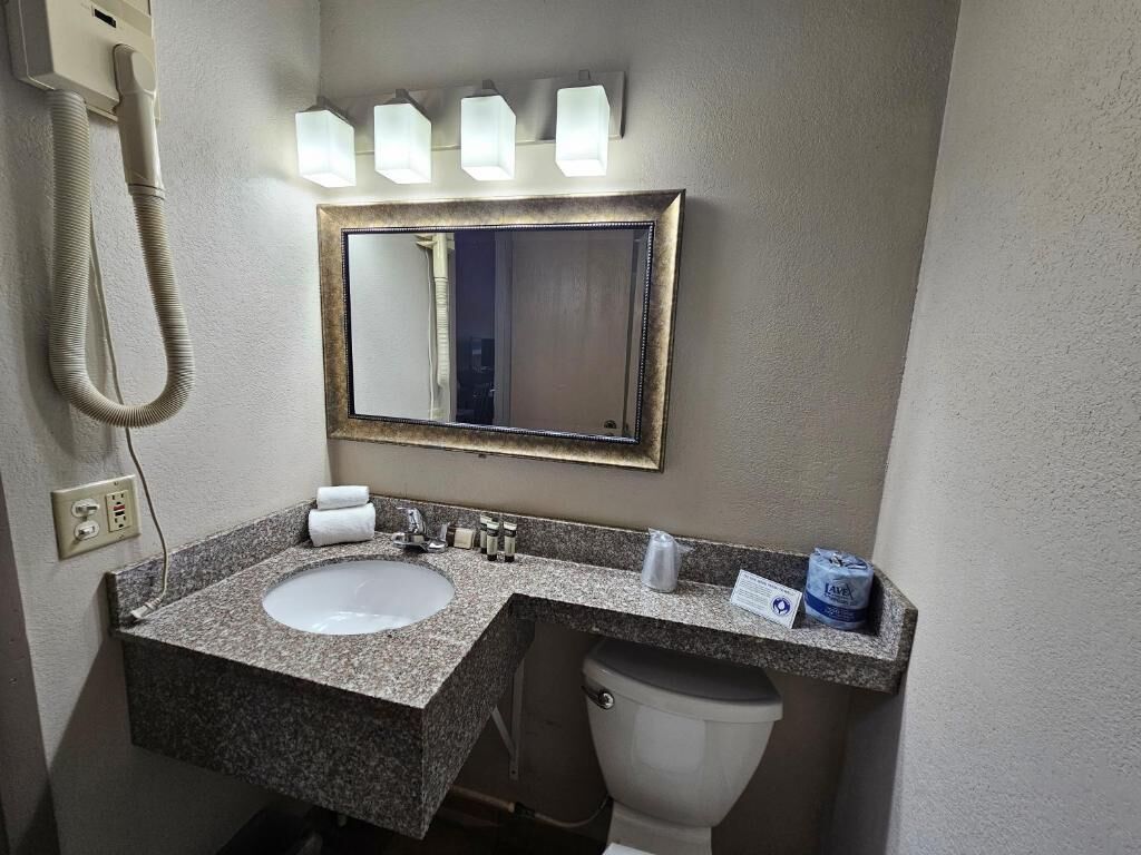 Un baño con lavamanos de encimera de granito, inodoro blanco, espejo con marco dorado y secador de pelo en la pared.