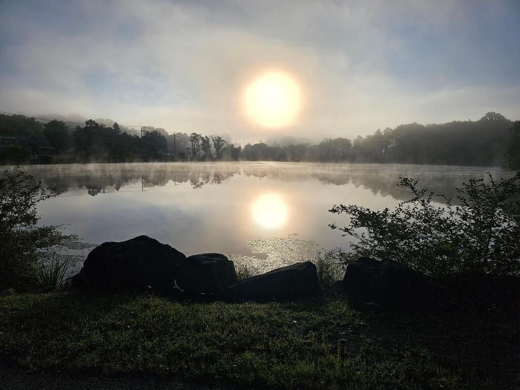 A calm lake with a clear reflection of the sun, surrounded by rocks and trees.