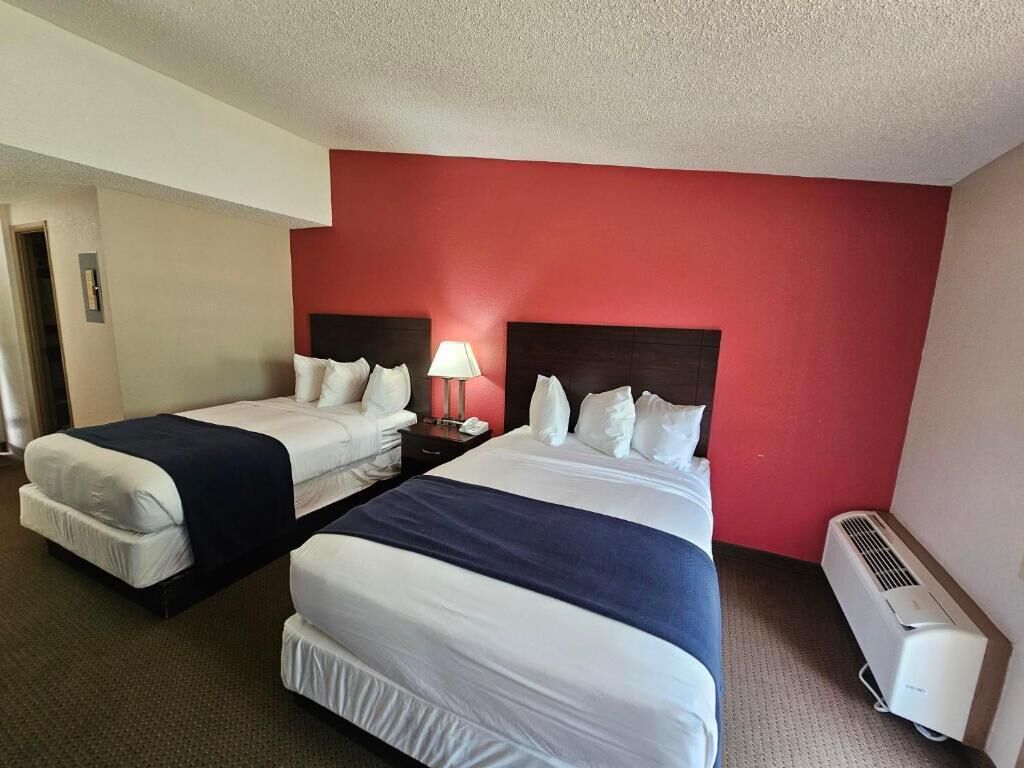 A hotel room with two beds, a red accent wall, a bedside table with a lamp, and an air conditioning unit.