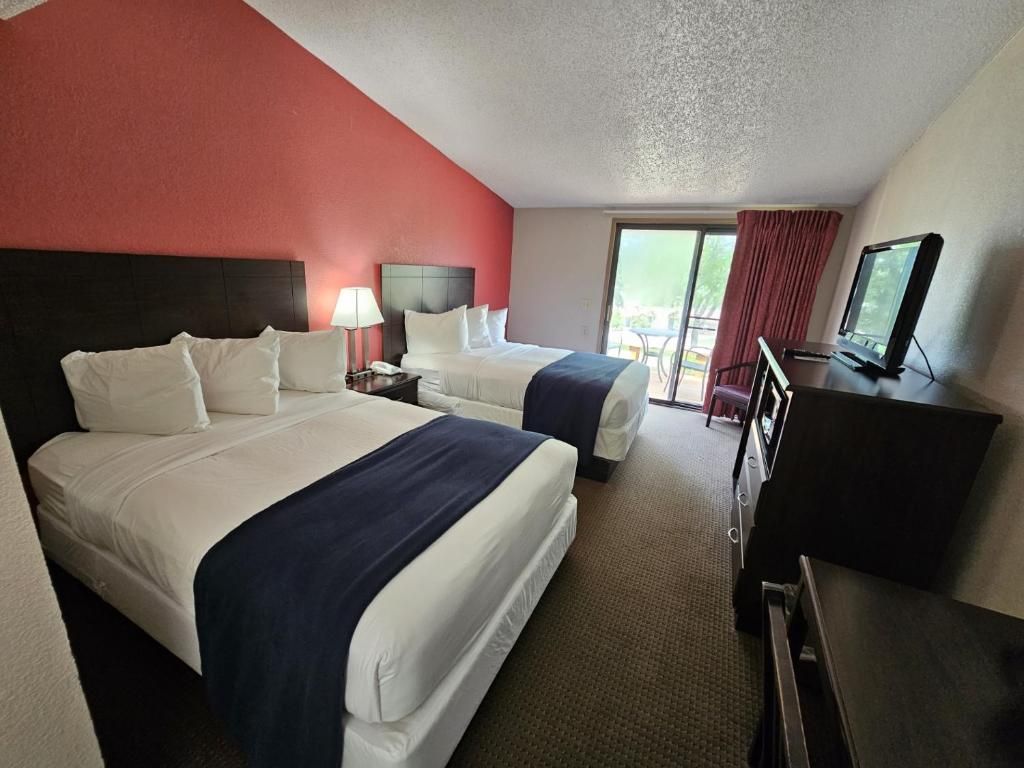 Habitación de hotel con dos camas, un escritorio, un televisor y un ventanal corredero que da a un espacio exterior.