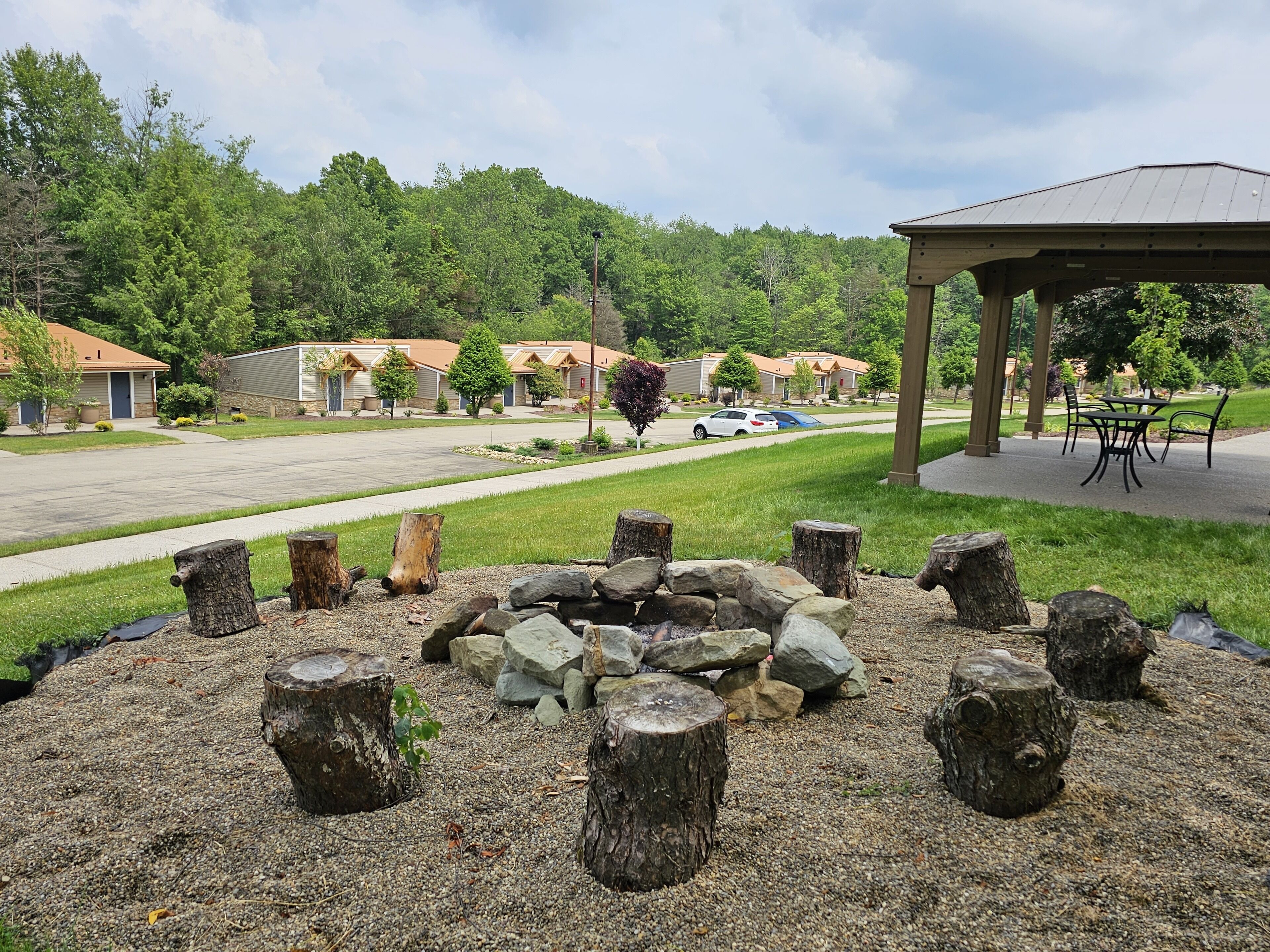 A residential area with a gazebo, a fire pit with logs and stones, and a row of houses with a green lawn and trees.