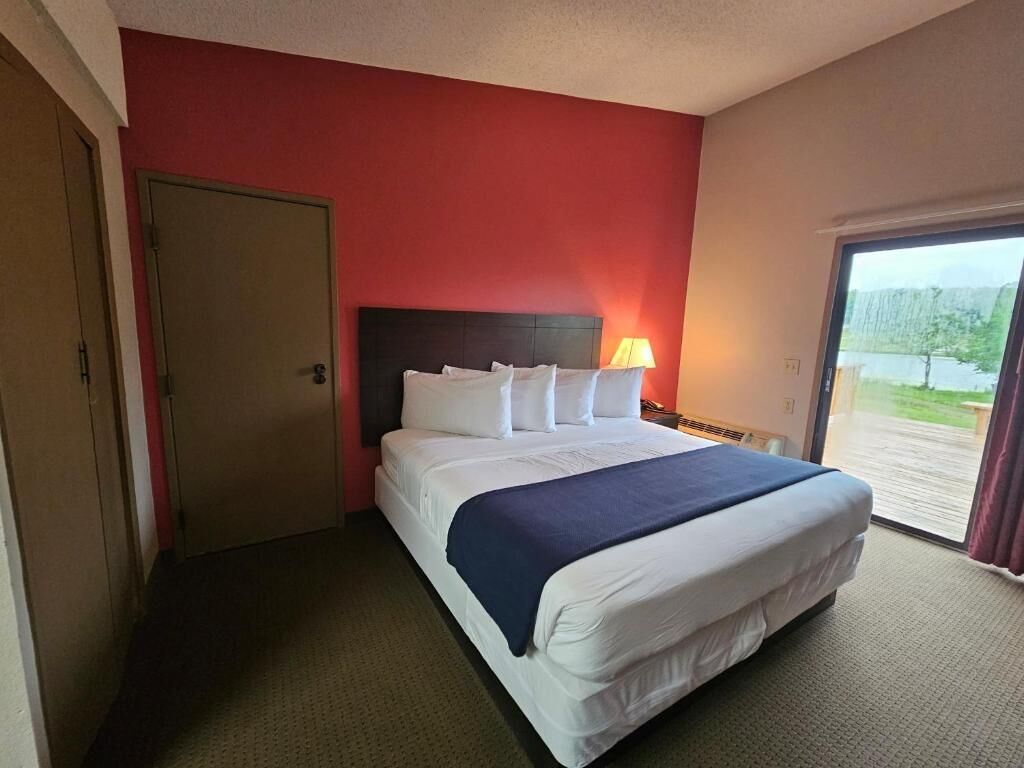 A hotel room with a bed, a nightstand with a lamp, a door, and a view of a natural landscape.