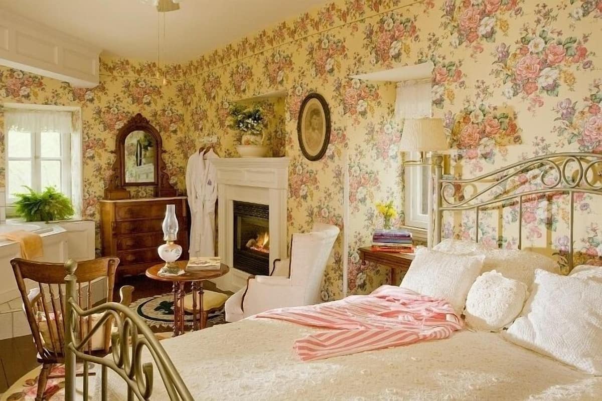 A bedroom with floral wallpaper, a fireplace, a bed with white linens, a wooden chair, and a side table with books.