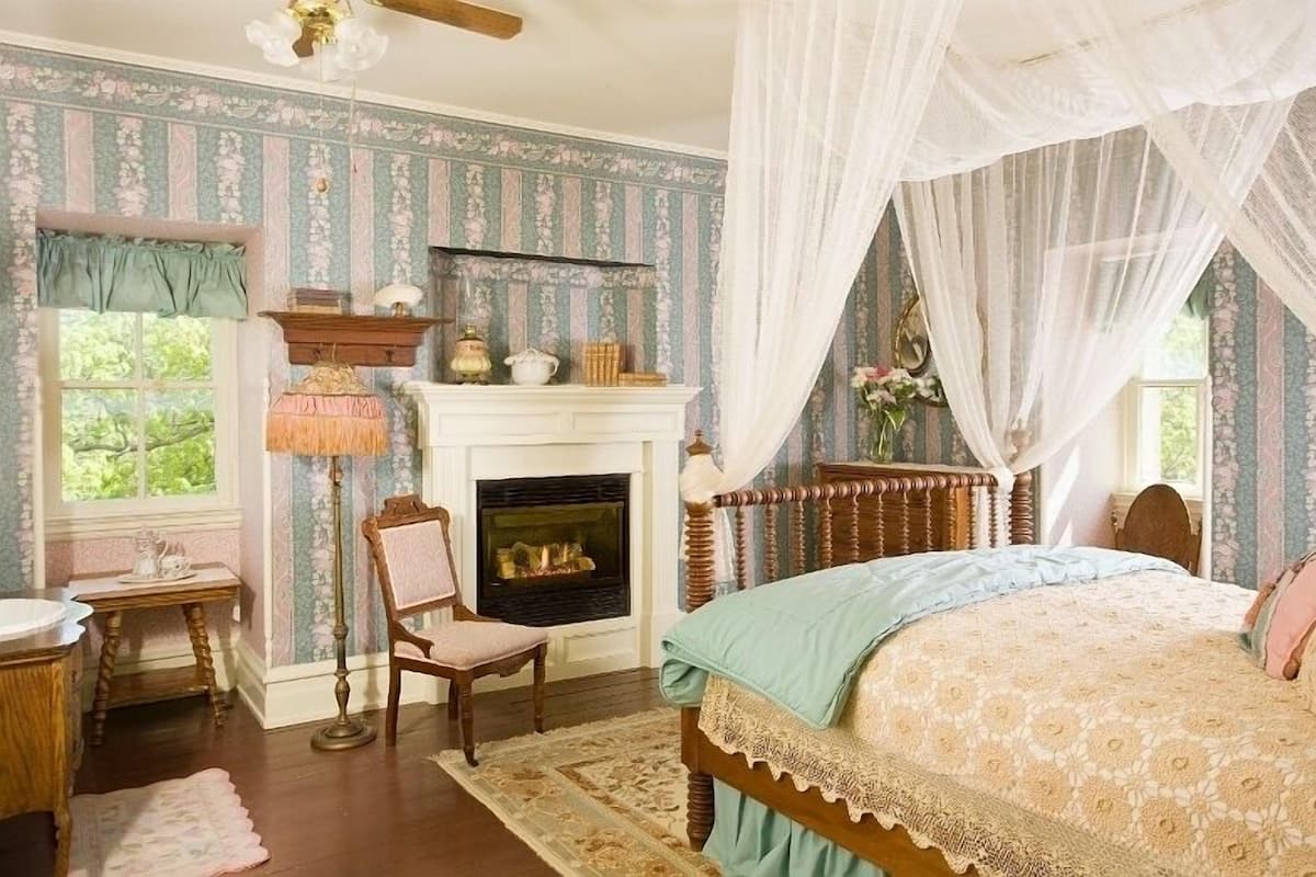 A bedroom with a fireplace, a bed with a canopy, a chair, and a table with a lamp.