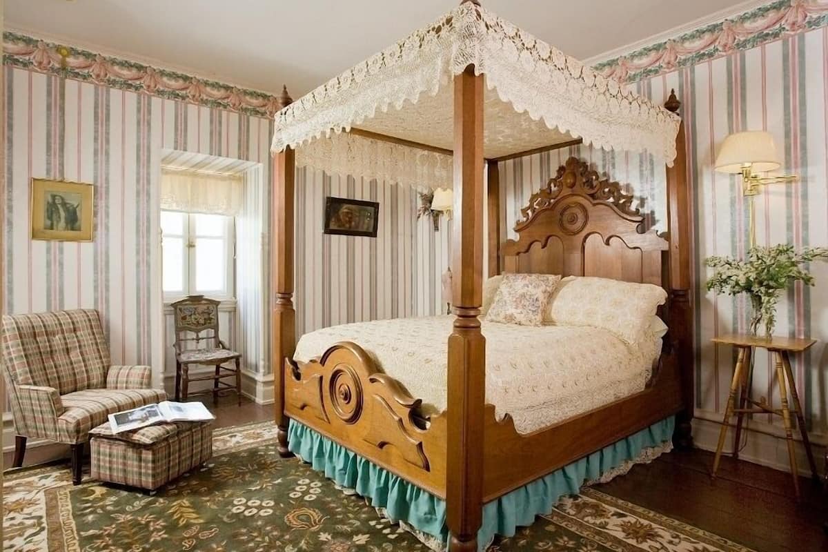 A four-poster canopy bed with a lace canopy, a plaid armchair, a small table, and a patterned rug.