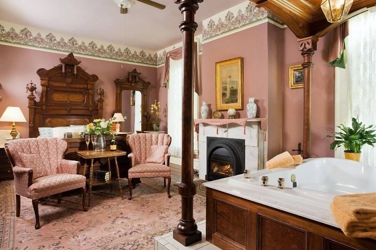 A traditional room with a large wooden bed, a fireplace, a sitting area with two chairs, and a table with a vase of flowers.
