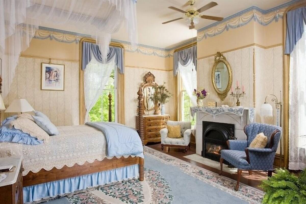 A bedroom with a four-poster bed, a fireplace, a blue armchair, and a chandelier.