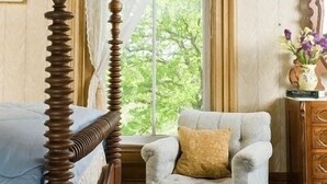 A bedroom with a four-poster bed, a tufted armchair, a wooden dresser, and a window with curtains.
