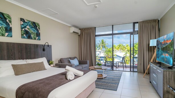 A modern hotel room with a large bed, a sofa, a TV, and a balcony with a view of palm trees. - Alivio Hotel Cairns (Cairns)