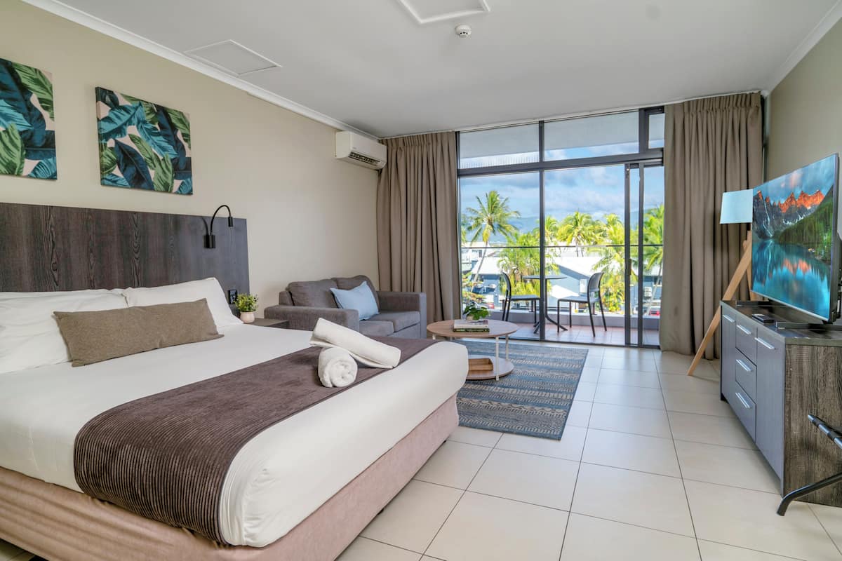 Una habitación de hotel moderna con una cama grande, un sofá, un televisor y un balcón con vistas a palmeras.