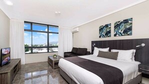 A modern hotel room with a large bed, a TV on a wooden stand, a small table, and a view of the city through large windows. - Alivio Hotel Cairns (Cairns)