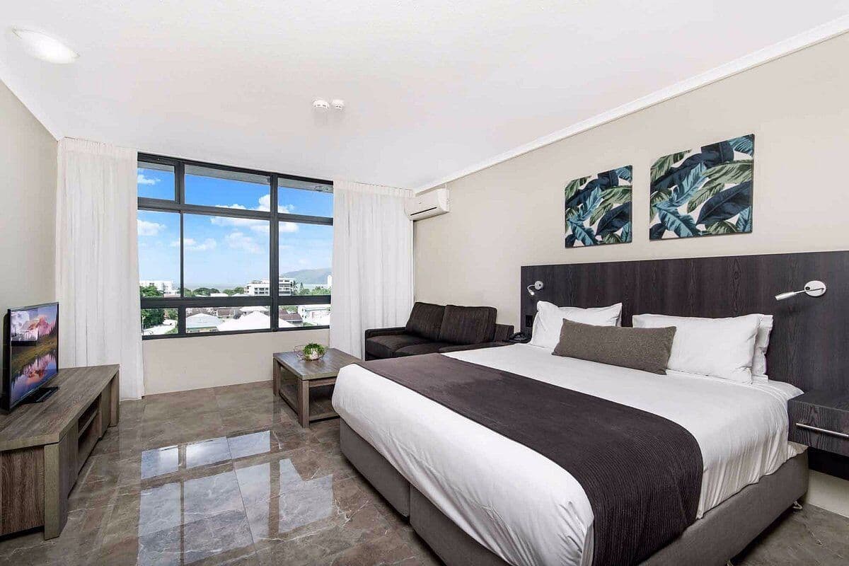 Habitación de hotel moderna con una cama grande, un televisor sobre un mueble de madera, una mesita pequeña y vistas a la ciudad a través de amplios ventanales.