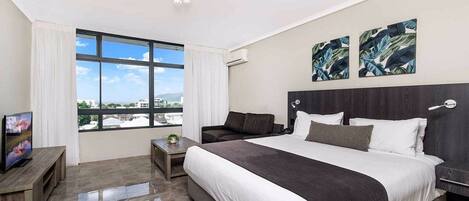 A modern hotel room with a large bed, a TV on a wooden stand, a small table, and a view of the city through large windows.