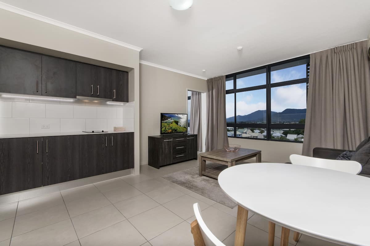 Una cocina moderna con armarios oscuros, una encimera blanca y un comedor con mesa y sillas blancas. Un televisor está sobre un mueble, y una ventana ofrece vistas a montañas.