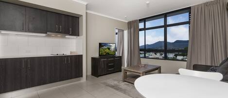 A modern kitchen with dark cabinets, a white countertop, and a dining area with a white table and chairs. A television is on a cabinet, and a window offers a view of mountains.