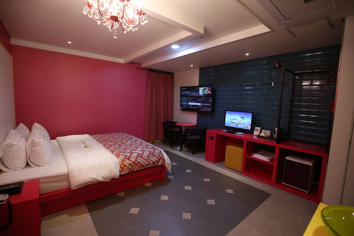 Moderni hotellihuone, jossa on punainen sänky, televisio, työpöytä ja pieni keittokomero.