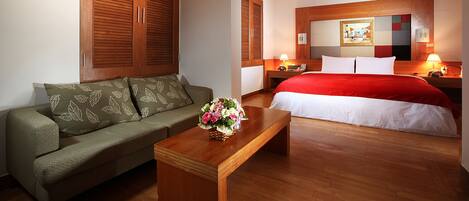 A hotel room with a wooden floor, a green sofa, a wooden coffee table with a floral arrangement, a bed with a red blanket, and a nightstand with a lamp.