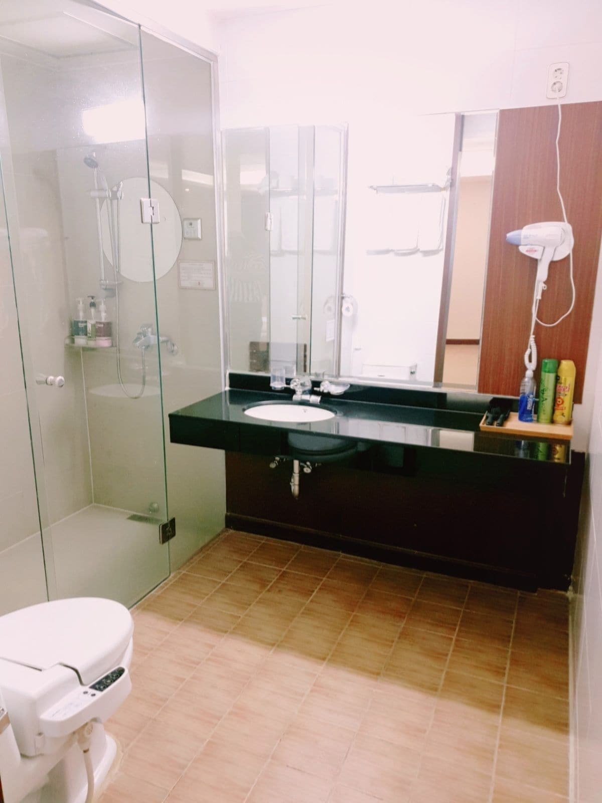 Un baño moderno con cabina de ducha de vidrio, inodoro blanco, lavamanos negro y un espejo encima, además de un secador de pelo en la pared.