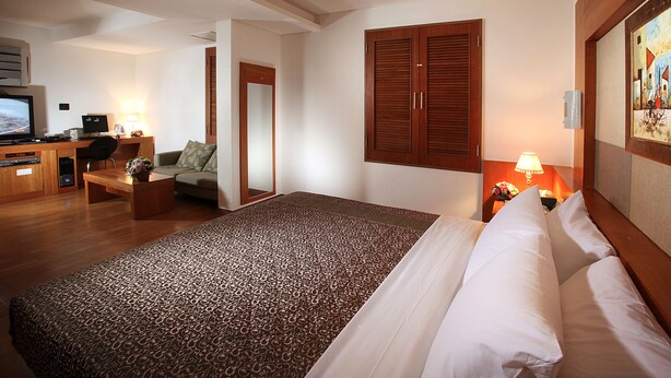 A hotel room with a large bed, a desk with a computer, a chair, and a small table with a lamp.