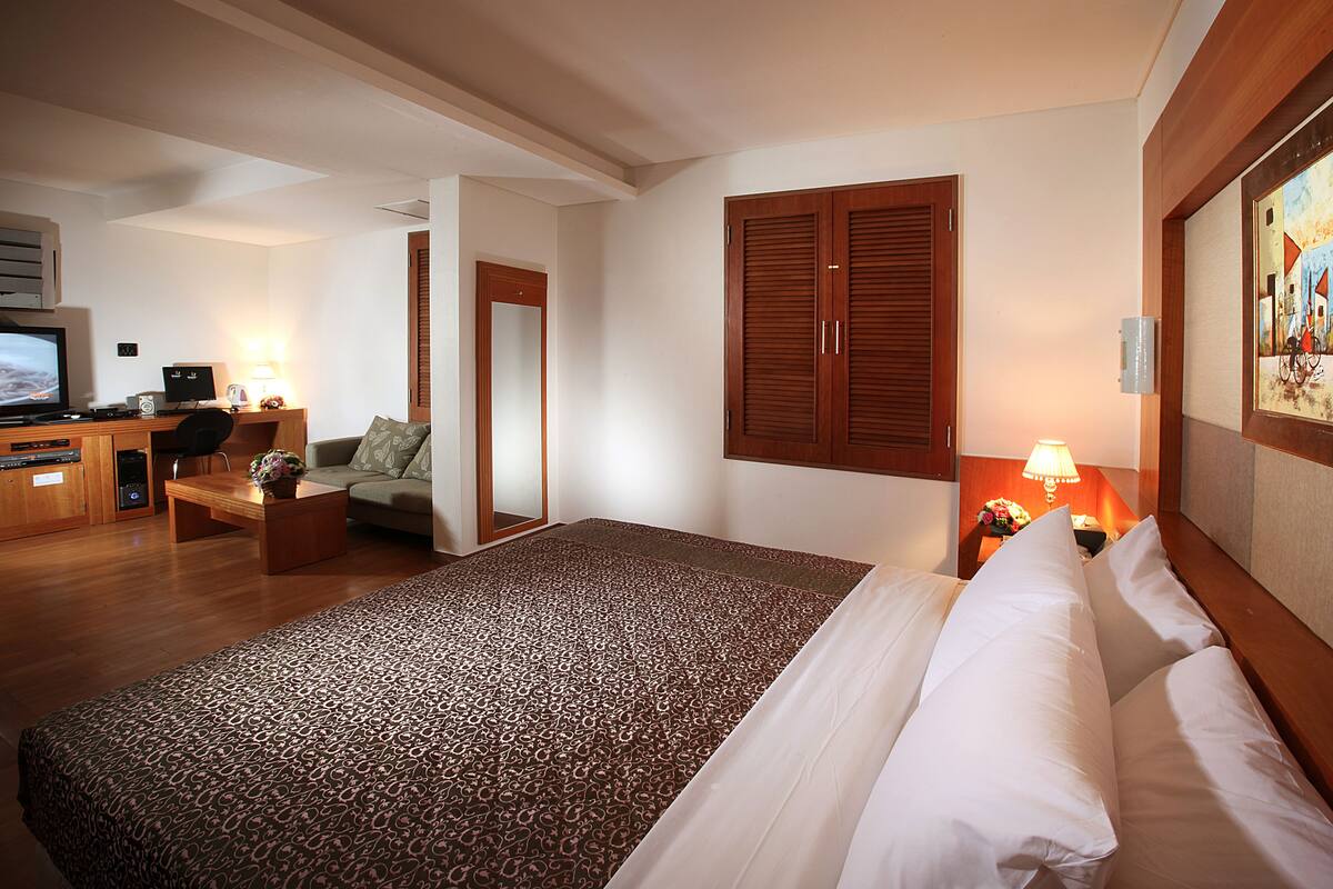 A hotel room with a large bed, a desk with a computer, a chair, and a small table with a lamp.