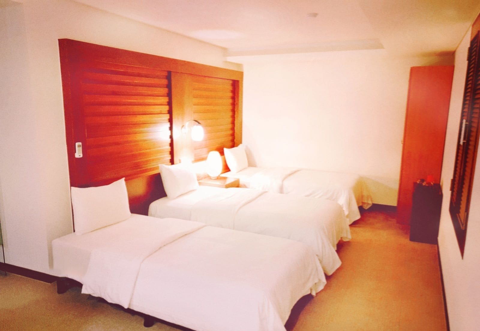 Habitación de hotel con dos camas, cabecera de madera, mesita de noche con lámpara y un cesto de basura pequeño.