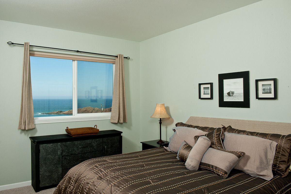 A bedroom with a bed, a nightstand, a lamp, a dresser, a window with curtains, and framed pictures on the wall.