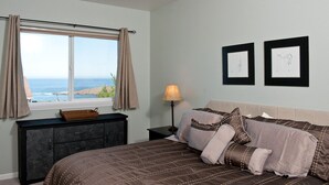 A bedroom with a bed, a nightstand, a lamp, a dresser, and a window with curtains. - Keystone Vacation Rentals-Ocean View Condos (Depoe Bay)